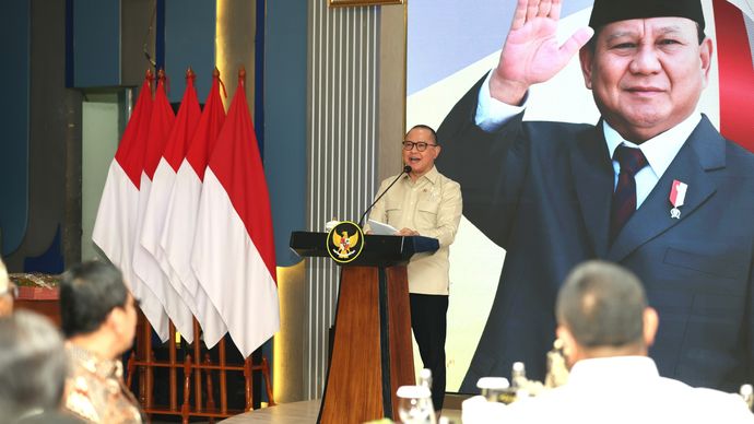 Menteri Pelindungan Pekerja Migran Indonesia (P2MI), Mukhtarudin, menyampaikan pidato dalam peringatan satu tahun masa jabatannya. Beliau menegaskan kembali komitmen negara untuk menghadirkan pelindungan bagi seluruh Pekerja Migran Indonesia. Latar belakang panggung menampilkan bendera merah putih dan foto besar Presiden Prabowo Subianto yang sedang melambaikan tangan, melambangkan dukungan penuh pemerintah.