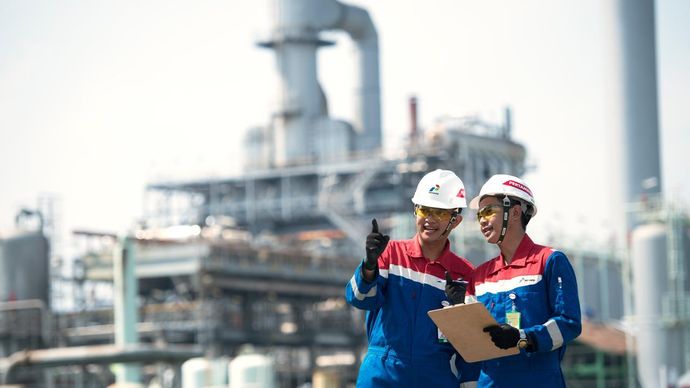 Dua pekerja PT Pertamina (Persero) mengenakan pakaian dinas lengkap dengan helm keselamatan dan papan catatan sedang memantau area operasional kilang, sejalan dengan komitmen perusahaan dalam mengembangkan bisnis energi hijau dan menurunkan emisi karbon untuk mendukung target Net Zero Emission 2060.